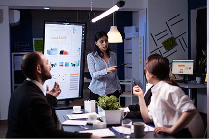 Workaholic businesswoman pointing financial strategy using monitor working overtime in company meeting office room. Diverse multi-ethnic teamwork overworked solving management statistics in evening