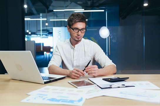 Portrait of a young handsome man, accountant, director, businessman, manager. Sitting in the office at the desk with glasses, working on a laptop, with a calculator and documents.