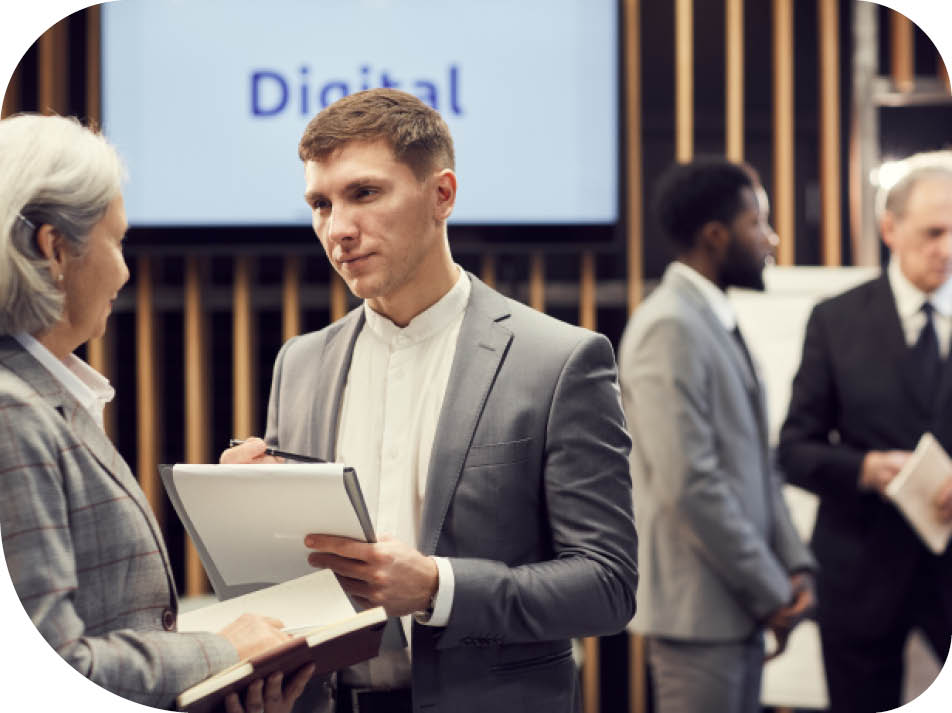 Serious handsome young journalist in suit making notes in clipboard while interviewing business forum participant during break