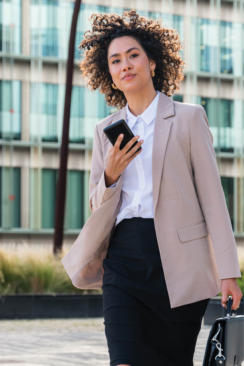 Beautiful hispanic businesswoman with elegant suit walking in the business centre- Adult female with business suit and holding mobile phone portrait outdoors