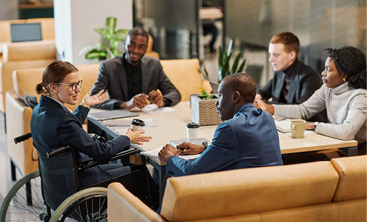 Portrait of successful businesswoman using wheelchair at meeting table and talking to diverse group of colleagues in modern office space